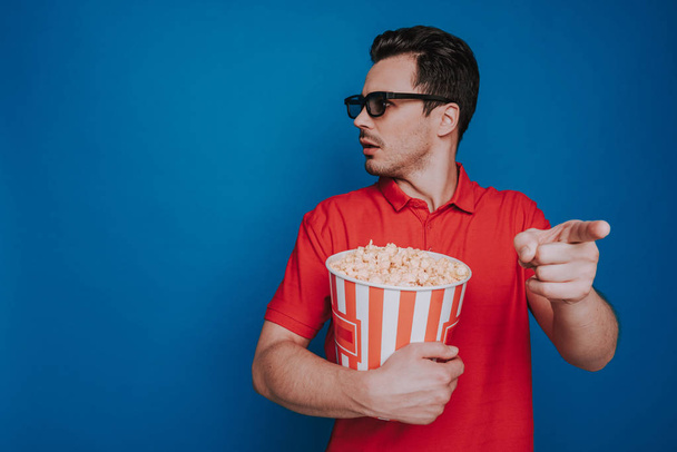 Waist up of surprised Caucasian man holding popcorn bucket on blue background - Foto, Imagen