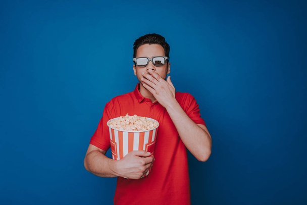 Waist up of surprised Caucasian man holding popcorn bucket on blue background - Фото, зображення
