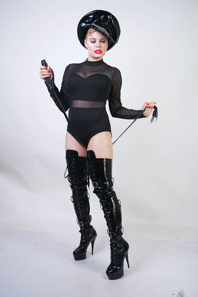 beautiful curvy strict mistress with red lips in black sexy bodysuit and patent leather fetish overknee boots holding a long leather whip on a white background in the Studio - Foto, Imagen