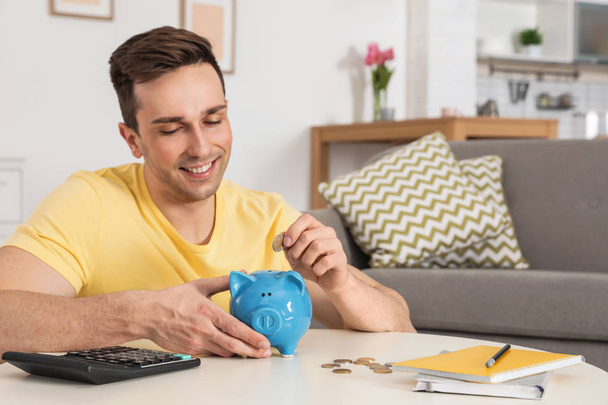 Happy man putting coin into piggy bank at table in living room. Saving money - Φωτογραφία, εικόνα