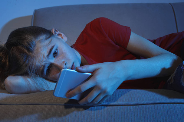 Upset teenage girl with smartphone lying on sofa in dark room. Danger of internet - Photo, Image
