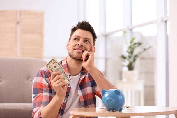 Man with piggy bank and money at home - Φωτογραφία, εικόνα
