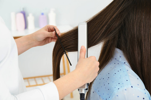 Hairdresser using modern flat iron to style client's hair in salon, closeup - Фото, зображення
