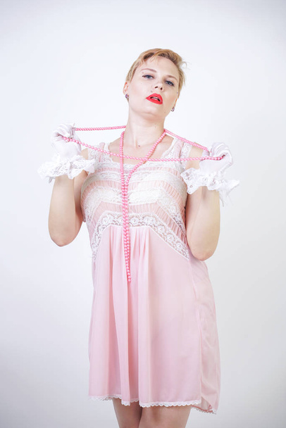 romantic plus size girl with short blonde hair in pink lace nightgown, white thin transparent gloves and pearl beads on white background in the Studio alone - Fotografie, Obrázek