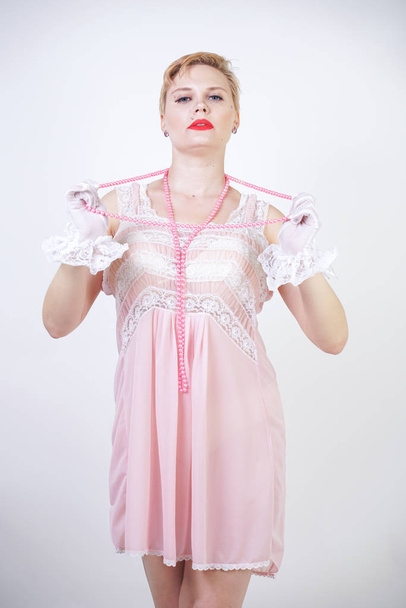 romantic plus size girl with short blonde hair in pink lace nightgown, white thin transparent gloves and pearl beads on white background in the Studio alone - Photo, Image