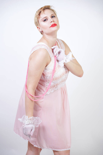 romantic plus size girl with short blonde hair in pink lace nightgown, white thin transparent gloves and pearl beads on white background in the Studio alone - Valokuva, kuva