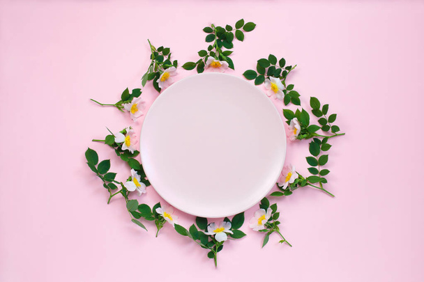 Colourful bright floral composition. Frame made of pink rose flowers and leafs around the pink plate on pastel pink background. Flat lay, copy space. - 写真・画像