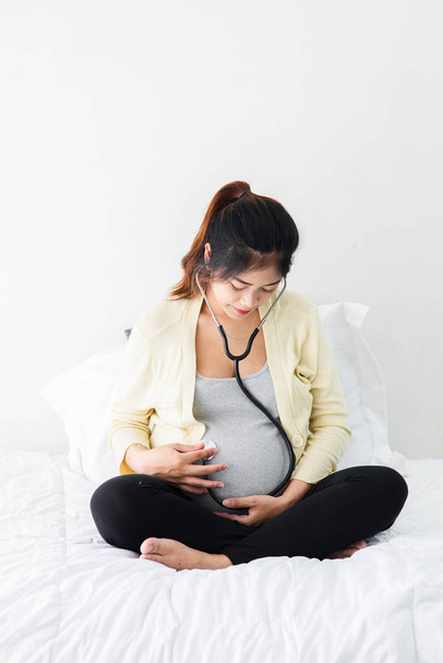 Asian pregnant woman using stethoscope listening her baby on bed - Fotoğraf, Görsel