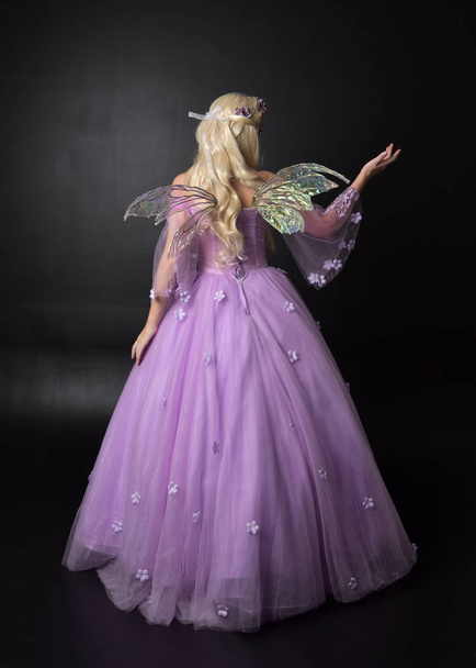 полноразмерный портрет блондинки в костюме фэнтези-феи, длинном фиолетовом бальном платье с крыльями феи, стоящей в позе спиной к камере на темном фоне студии
. - Фото, изображение