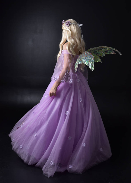teljes hosszúságú portréja egy szőke lány visel fantasy tündér ihlette jelmez, hosszú lila golyó ruháját tündér szárnyak, álló pózol vissza a kamerát egy sötét stúdió háttérben. - Fotó, kép