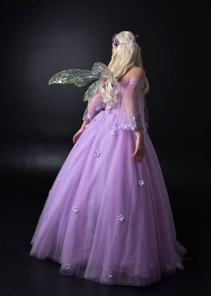 Pełna długość Portret blond dziewczyna ubrana w fantazyjne bajki inspirowane kostium, długa fioletowa suknia balowa z bajki skrzydła, stojący poza z powrotem do aparatu na ciemnym tle Studio. - Zdjęcie, obraz