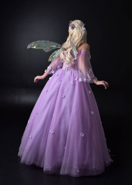 портрет блондинки в фентезі фея, натхненний костюм, довга фіолетова кулькова сукня з казковими крилами, стояча поза спиною до камери на темному студійному фоні
. - Фото, зображення