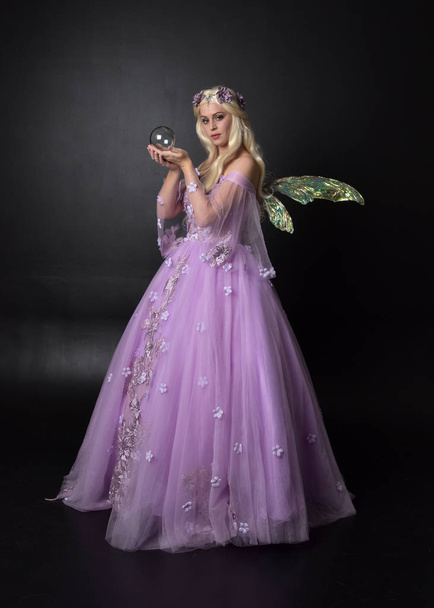 teljes hosszúságú portréja egy szőke lány visel fantasy tündér ihlette jelmez, hosszú lila golyó ruháját tündér szárnyak, ül jelentenek a sötét stúdió háttér. - Fotó, kép