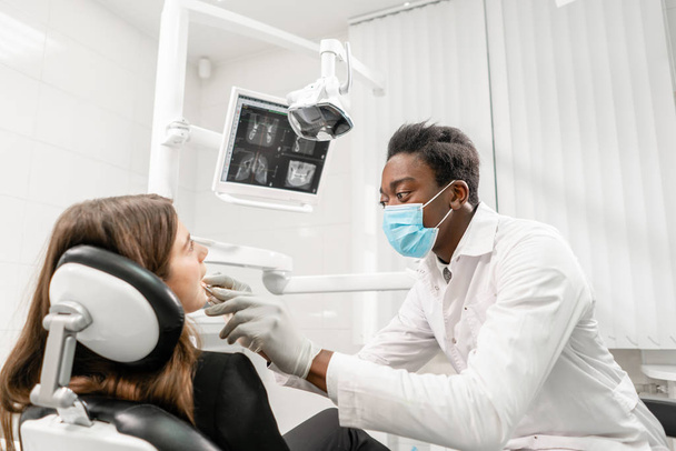 Doctor treats teeth. Young African male dentist with a patient. Woman in the dentist chair at dental clinic. Medicine, health, stomatology concept. dentist treating a patient. - Foto, Imagem