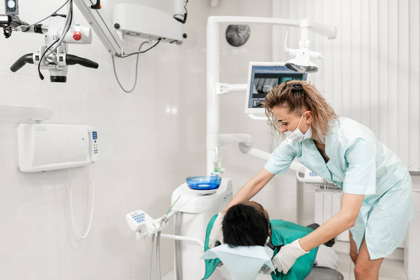 Γυναίκα οδοντίατρος χρησιμοποιεί μηχάνημα ακτίνων χ, ασθενής ξαπλωμένος στην καρέκλα στην οδοντιατρική. Νέοι αφροαμερικάνων άνδρες με χαλασμένα δόντια. Ιατρική, υγεία, Στοματολογία έννοια. - Φωτογραφία, εικόνα