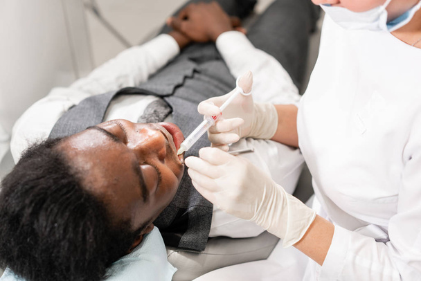 Jeune femme dentiste traitant les canaux radiculaires dans la clinique dentaire. Jeune homme afro-américain avec de mauvaises dents couché sur une chaise de dentiste à bouche ouverte
. - Photo, image