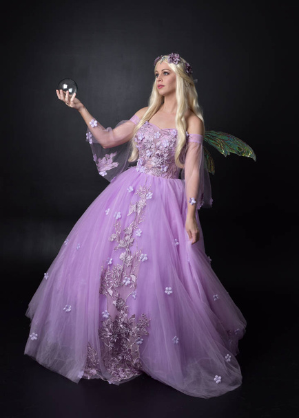 Pełna długość Portret blond dziewczyna ubrana w fantazyjne bajki inspirowane kostium, długa fioletowa suknia balowa z bajki skrzydła, siedząc pozować na ciemnym tle Studio. - Zdjęcie, obraz