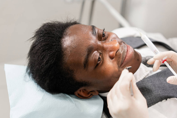 Молодая женщина-дантист лечит корневые каналы в стоматологической клинике. Молодой афроамериканец с плохими зубами лежит на стуле стоматолога с открытым ртом
. - Фото, изображение