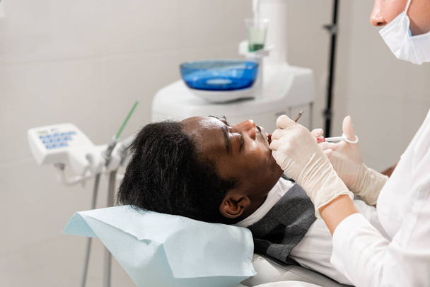 Молода жінка-стоматолог лікуванні кореневих каналів в стоматологічній клініці. Молоді афроамериканець чоловіки з поганим зубами лежачи на стоматолога голова з відкритим ротом. - Фото, зображення
