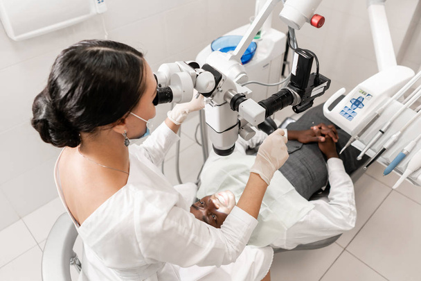 Молодая женщина-дантист лечит корневые каналы с помощью микроскопа в стоматологической клинике. Пациент лежит на стоматологическом стуле с открытым ртом. Медицина, стоматология и здравоохранение концепция. Стоматологическое оборудование
 - Фото, изображение