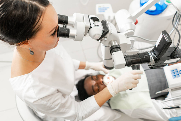 Microscope d'équipement moderne dans le bureau dentaire. Jeune femme dentiste traitant les canaux radiculaires. Homme patient allongé sur une chaise de dentiste à bouche ouverte. Concept de médecine, dentisterie et soins de santé
. - Photo, image