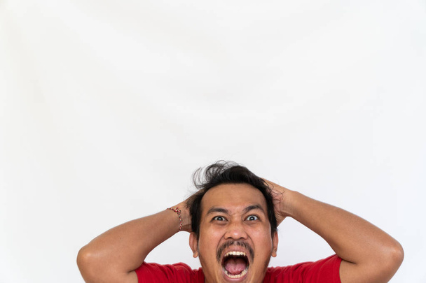Enraged furious asian man open his mouth and screaming in anger and stress, pulling his hair out with his hand, stand isolated on white background and make grimace closed eyes. Space for text - Photo, Image