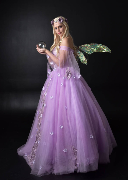 Ganzkörperporträt eines blonden Mädchens, das ein fantasievolles, von Feen inspiriertes Kostüm trägt, ein langes lila Ballkleid mit Feenflügeln, das auf dunklem Studiohintergrund posiert. - Foto, Bild