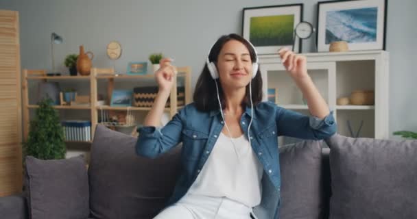 Joyful woman listening to music moving hands and head sitting on sofa at home - Video, Çekim