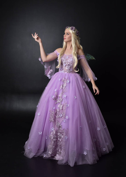 full length Portret van een blonde meisje het dragen van een fantasie fee geïnspireerd kostuum, lange paarse baljurk met sprookjes vleugels, zittend poseren op een donkere studio achtergrond. - Foto, afbeelding