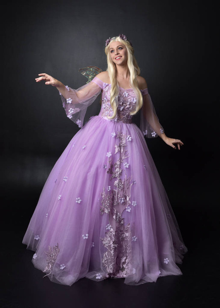 full length portrait of a blonde girl wearing a fantasy fairy inspired costume,  long purple ball gown with fairy wings,   sitting pose  on a dark studio background. - Photo, Image