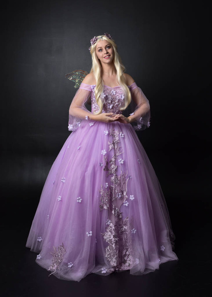 Pełna długość Portret blond dziewczyna ubrana w fantazyjne bajki inspirowane kostium, długa fioletowa suknia balowa z bajki skrzydła, siedząc pozować na ciemnym tle Studio. - Zdjęcie, obraz