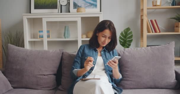 Beautiful girl writing in notebook looking at smartphone screen sitting at home - Filmagem, Vídeo