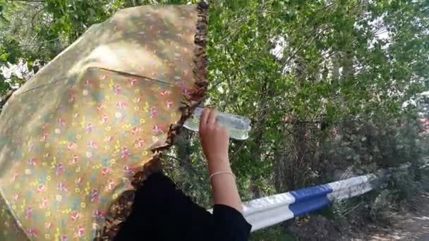 una chica en un paraguas camina por la carretera en un clima cálido y soleado para saciar su sed bebiendo una botella de agua
 - Metraje, vídeo