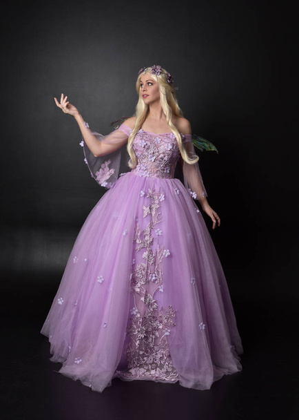 full length portrait of a blonde girl wearing a fantasy fairy inspired costume,  long purple ball gown with fairy wings,   sitting pose  on a dark studio background. - Fotó, kép