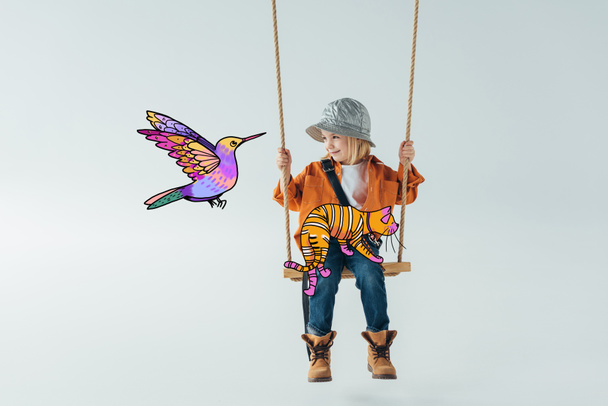 cute kid in jeans and orange shirt sitting on swing with fantasy cat on knees and looking at colorful bird illustration on grey background - Photo, Image