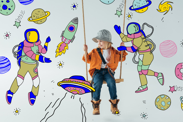 cute kid in jeans and orange shirt sitting on swing and looking at fairy space with astronauts illustration on grey background - Photo, Image