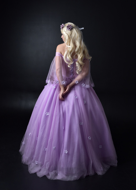 full length portrait of a blonde girl wearing a fantasy fairy inspired costume,  long purple ball gown with fairy wings,   standing pose with back to the camera on a dark studio background. - 写真・画像