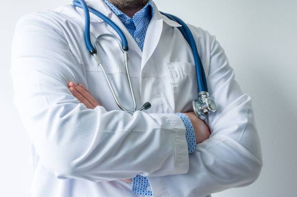 Photo of body of doctor in white lab coat with stethoscope on his neck in half-turn with folded hands against white wall in hospital with light source on left. Concept image of  medical worker  - Foto, afbeelding