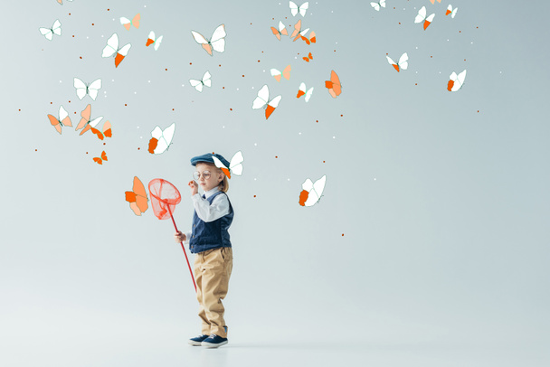 χαριτωμένο και ξανθιά παιδί σε ρετρό γιλέκο και καπάκι κρατώντας πεταλούδα δίχτυ σε γκρι φόντο με πεταλούδες νεράιδα - Φωτογραφία, εικόνα
