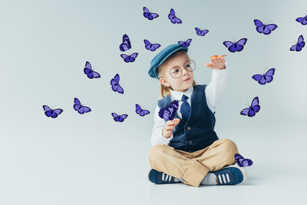 adorable kid sitting on floor with crossed legs among fairy purple butterflies on grey background  - Photo, Image
