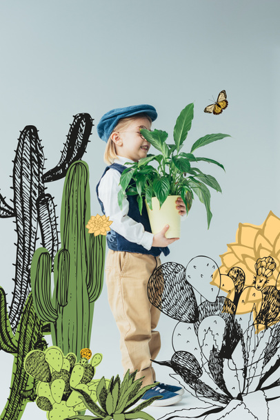 adorable kid in retro vest and cap holding plant in flowerpot on grey background with fairy cacti illustration - Photo, Image