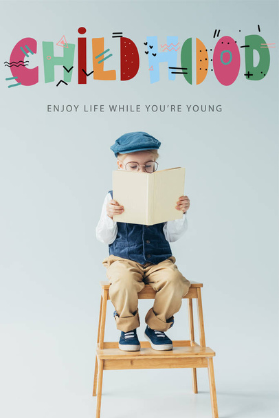 carino bambino seduto su scale di legno e libro di lettura su sfondo grigio con lettering infanzia
 - Foto, immagini
