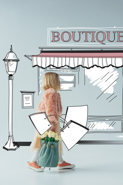 side view of cute kid with white tulips and shopping bags looking at fairy boutique on street on grey background - Photo, Image