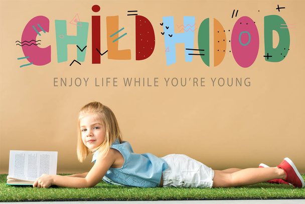 carino bambino sdraiato su tappeto di erba e tenendo libro su sfondo beige con illustrazione infantile
  - Foto, immagini