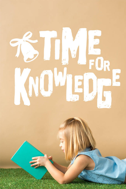 smiling and cute child lying on grass rug and holding book on beige background with white time for knowledge lettering - Fotó, kép