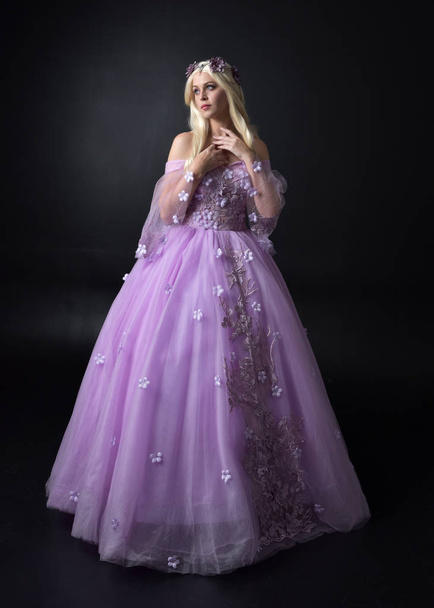 full length portrait of a blonde girl wearing a fantasy fairy inspired costume,  long purple ball gown with fairy wings,   standing pose on a dark studio background. - Photo, Image