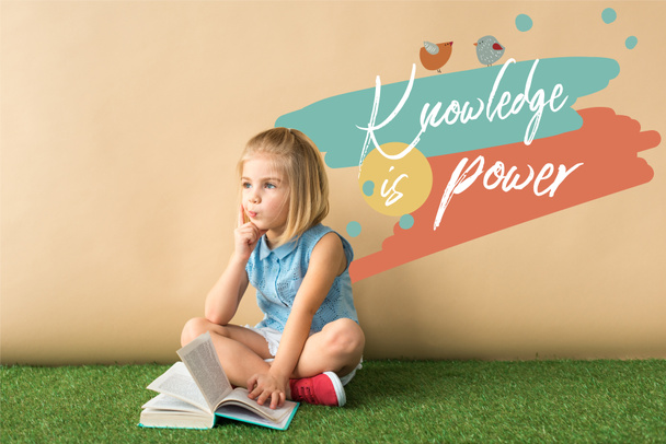 thoughtful cute child sitting with crossed legs on grass rug and holding book on beige background with knowledge is power lettering  - Photo, Image