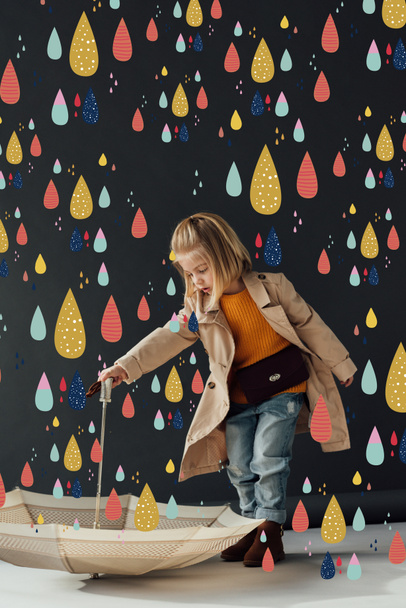 serious child in trench coat and jeans holding umbrella under fairy colorful rain  - Photo, Image