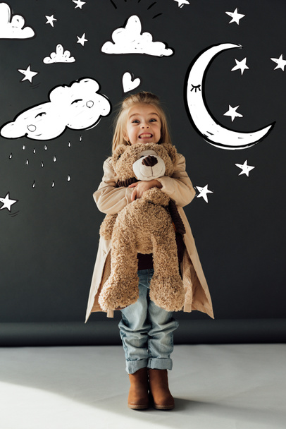 happy and cute child in trench coat and jeans hugging teddy bear on black background with magic moon, stars and rainy cloud illustration - Photo, Image