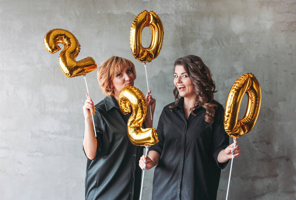 Dos mujeres jóvenes con vestidos negros sosteniendo los globos de números 2020 sobre fondo de pared de hormigón gris. Fiesta de Año Nuevo con amigos, corporativo
 - Foto, Imagen